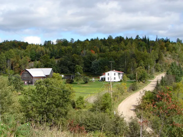 Chemin de terre qui monte vers une ferme et une maison blanche entourées de forêt à Boileau