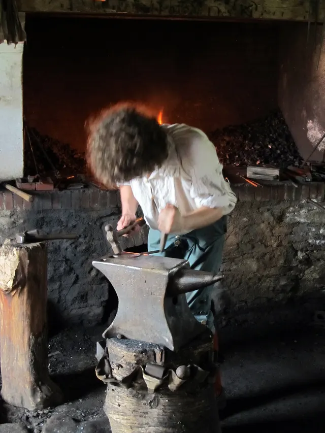 Forgeron qui martèle une pièce sur l'enclume, devant la forge